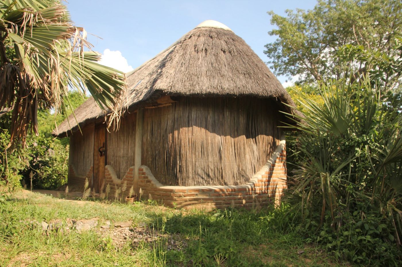 Strohdachbedeckte Gästehütte mit Schilfwänden und Backsteinfundament, zwischen Palmen, im Liwonde Safari Camp in Liwonde.